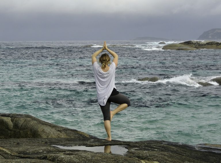 Woman balancing on one leg in tree pose on a rock overlooking the ocean