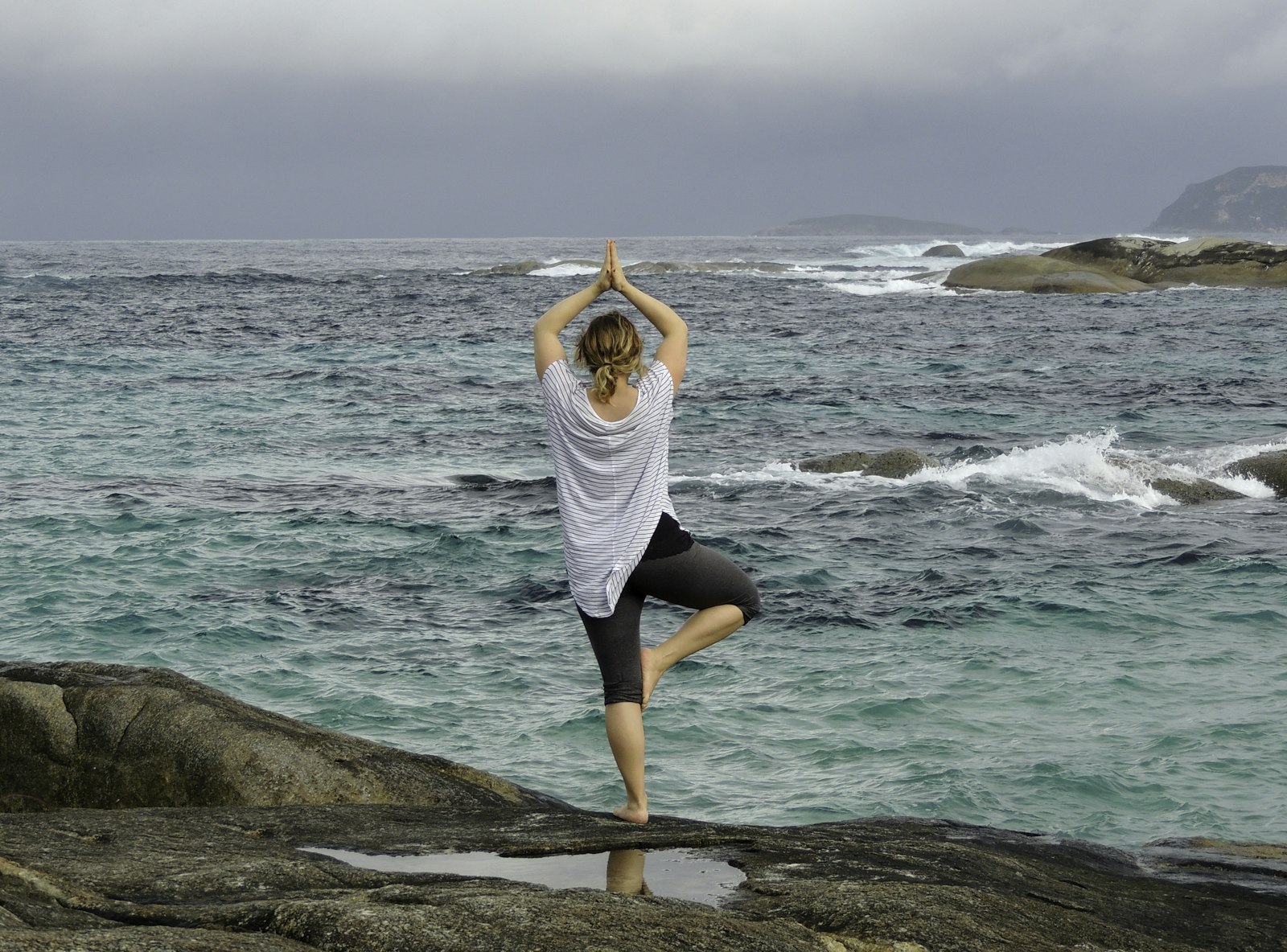 Woman balancing on one leg in tree pose on a rock overlooking the ocean