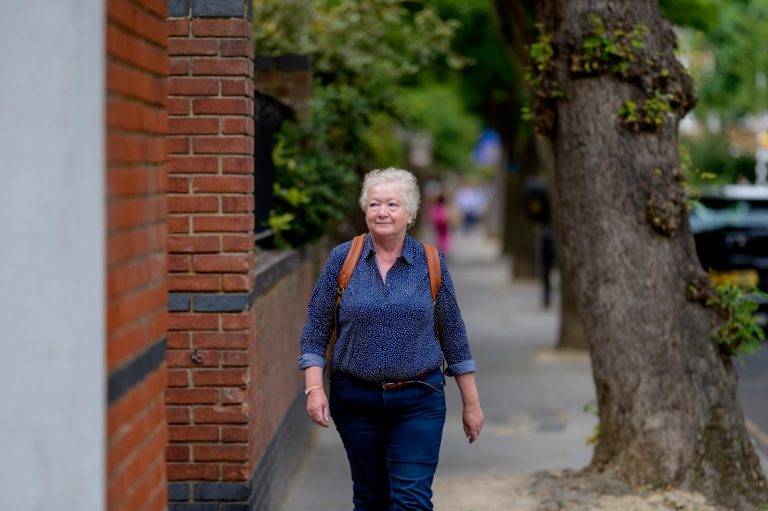 Older woman walking down a leafy sidewalk with a backpack