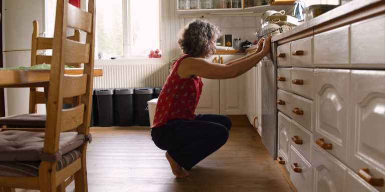Woman with grey hair squatting down to a low cupboard in her kitchen