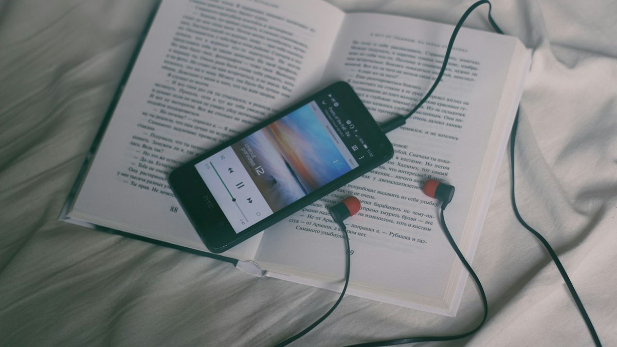 A phone and earbuds resting on an open book in bed
