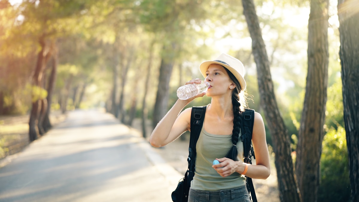 Woman in a sun hat drinking from a water bottle on a tree-lined path